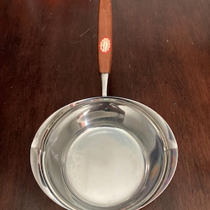 以下が含まれることがあります： 木製のハンドルが付いたステンレス製のソースパン。ハンドルは明るい茶色で、小さな丸いステッカーが付いています。鍋は丸く、光沢のある反射面です。 キッチン用品。