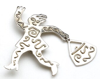 Puede incluir: Broche plateada con una figura estilizada de una persona corriendo con un bolso. La figura y el bolso están decorados con intrincados patrones en espiral.