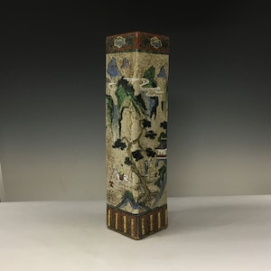 May include: A white ceramic vase with a square shape and a painted design of a landscape with trees, mountains, and figures. The vase has a red, yellow, and blue border.