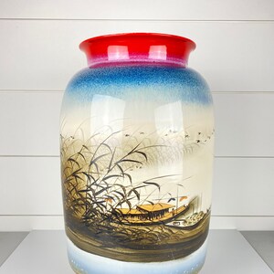 May include: A large ceramic vase with a red rim, transitioning to blue and white. The vase features a painted scene of a boat, reeds, and birds. The vase is approximately 14 inches tall.