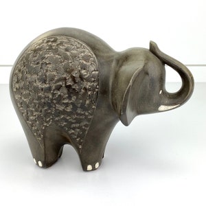 Puede incluir: Figura de elefante gris oscuro con la espalda texturizada. El elefante tiene una trompa levantada y pequeños detalles blancos en las patas y la trompa. La figura es un objeto decorativo, probablemente para una estantería o mesa.