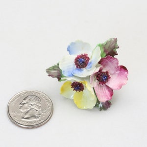 Peut inclure: Une broche en porcelaine en forme de fleurs avec trois fleurs dans des tons de bleu, jaune et rose. Les fleurs ont des centres bordeaux et des feuilles vertes.