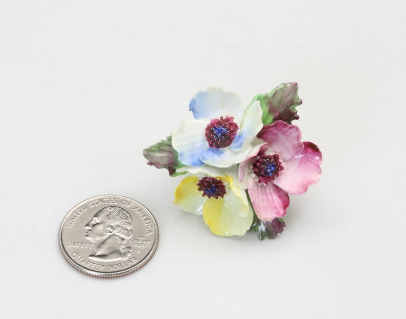 Peut inclure: Une broche en porcelaine en forme de fleurs avec trois fleurs dans des tons de bleu, jaune et rose. Les fleurs ont des centres bordeaux et des feuilles vertes.