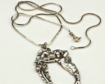 May include: A silver pendant necklace with an Art Nouveau design. The pendant shows two figures in an oval frame, with detailed floral and flowing lines. The chain is a fine box chain style.