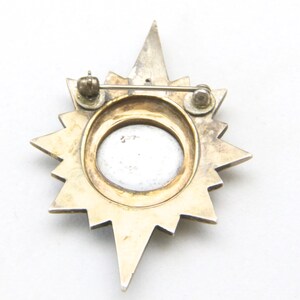 May include: A vintage starburst brooch with a silver-tone and gold-tone finish. The brooch features a central oval design and a pin closure on the back. The starburst design has pointed rays extending outwards.