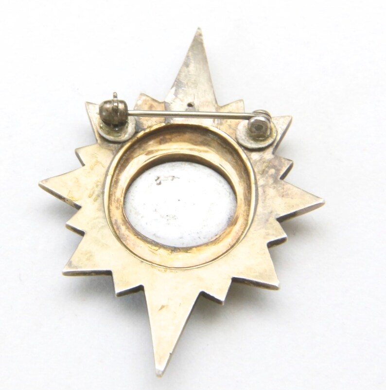 May include: A vintage starburst brooch with a silver-tone and gold-tone finish. The brooch features a central oval design and a pin closure on the back. The starburst design has pointed rays extending outwards.