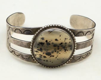 May include: Silver cuff bracelet with a large, round, speckled stone centre. The bracelet has three horizontal bands and decorative stamped details. The stone is surrounded by a beaded border.