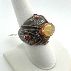 May include: A silver ring with a textured, dark gray band and gold accents. The ring features a large, oval, light yellow gemstone and smaller red gemstones. The ring is displayed on a white finger form.