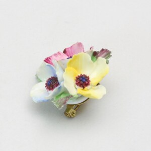 Peut inclure: Une broche en porcelaine repr&eacute;sentant un bouquet de trois fleurs aux nuances de rose, jaune et bleu. Les fleurs ont des p&eacute;tales et des centres d&eacute;taill&eacute;s, et la broche a une &eacute;pingle dor&eacute;e.
