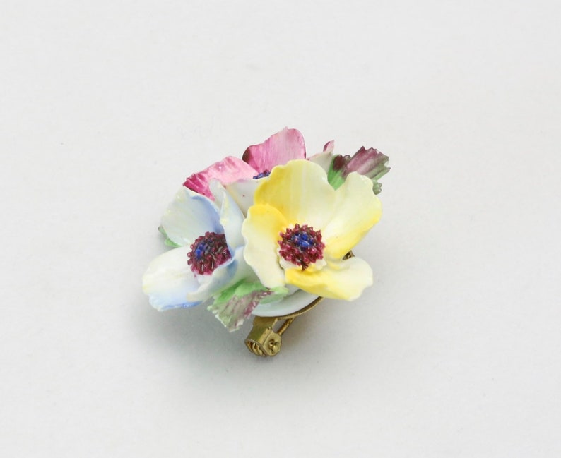 Peut inclure: Une broche en porcelaine repr&eacute;sentant un bouquet de trois fleurs aux nuances de rose, jaune et bleu. Les fleurs ont des p&eacute;tales et des centres d&eacute;taill&eacute;s, et la broche a une &eacute;pingle dor&eacute;e.