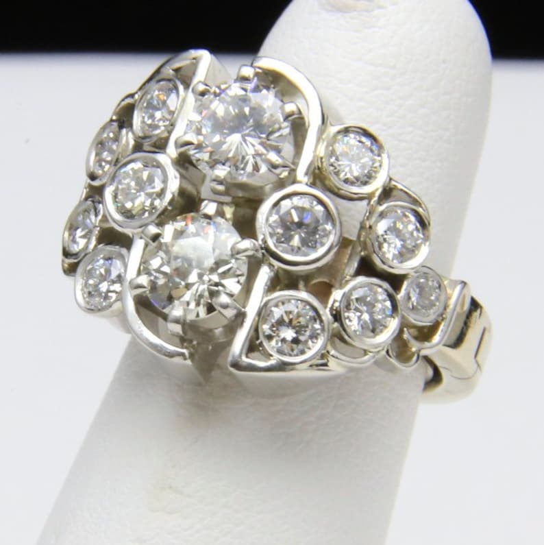 Puede incluir: Un anillo de oro blanco con m&uacute;ltiples diamantes redondos. El anillo tiene un dise&ntilde;o de racimo con diamantes m&aacute;s grandes en el centro y diamantes m&aacute;s peque&ntilde;os alrededor. El anillo se muestra en un dedo.