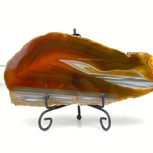 May include: A polished agate slice, showcasing bands of orange, brown, white, and gray hues. The stone is displayed on a black metal stand with a three-legged design. The agate's natural patterns create a unique decorative piece.
