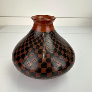 Op de afbeelding: Een ronde keramische vaas met een gladde, effen terracotta-kleurige hals en een brede, bolvormige buik. De buik is voorzien van een herhalend geometrisch patroon van zwarte vierkanten en rechthoeken, overlapt door dunne, kruisende terracotta-kleurige lijnen.