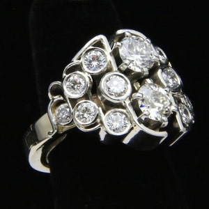 Puede incluir: Un anillo de plata con m&uacute;ltiples piedras preciosas redondas y transparentes. El anillo presenta un grupo de piedras m&aacute;s grandes rodeadas de otras m&aacute;s peque&ntilde;as, en un dise&ntilde;o calado &uacute;nico. El anillo se muestra sobre un fondo negro.
