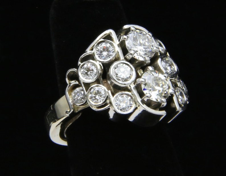 Puede incluir: Un anillo de plata con m&uacute;ltiples piedras preciosas redondas y transparentes. El anillo presenta un grupo de piedras m&aacute;s grandes rodeadas de otras m&aacute;s peque&ntilde;as, en un dise&ntilde;o calado &uacute;nico. El anillo se muestra sobre un fondo negro.