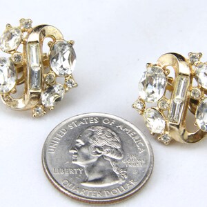 May include: A pair of gold tone clip-on earrings with clear rhinestone accents. The earrings are shaped like a figure eight with a row of baguette-shaped rhinestones in the center.