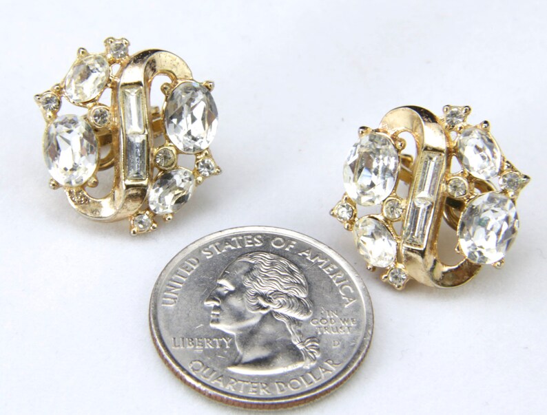 May include: A pair of gold tone clip-on earrings with clear rhinestone accents. The earrings are shaped like a figure eight with a row of baguette-shaped rhinestones in the center.