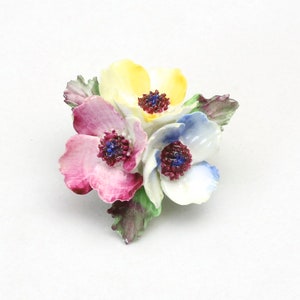 Peut inclure: Une broche en porcelaine en forme de fleurs avec trois fleurs dans des tons de rose, jaune et bleu. Les fleurs ont des centres blancs avec des accents rouge fonc&eacute;.