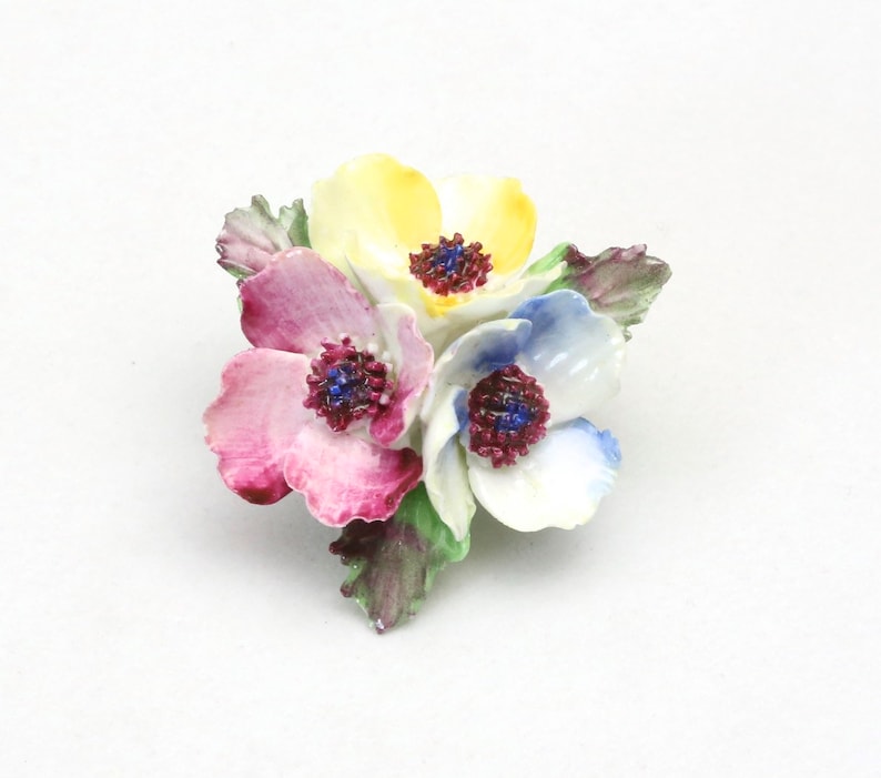 Peut inclure: Une broche en porcelaine en forme de fleurs avec trois fleurs dans des tons de rose, jaune et bleu. Les fleurs ont des centres blancs avec des accents rouge fonc&eacute;.