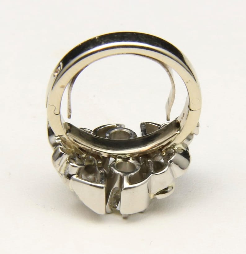 Puede incluir: Un anillo plateado con un grupo de peque&ntilde;as piedras transparentes. La banda del anillo es de color plata brillante, y el engaste alrededor de las piedras tiene un dise&ntilde;o geom&eacute;trico detallado. El anillo se muestra desde un &aacute;ngulo, resaltando su estructura tridimensional.