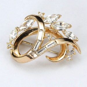 May include: A gold-toned brooch with a swirling design and clear rhinestones. The brooch features a variety of rhinestone shapes, including marquise and baguette.