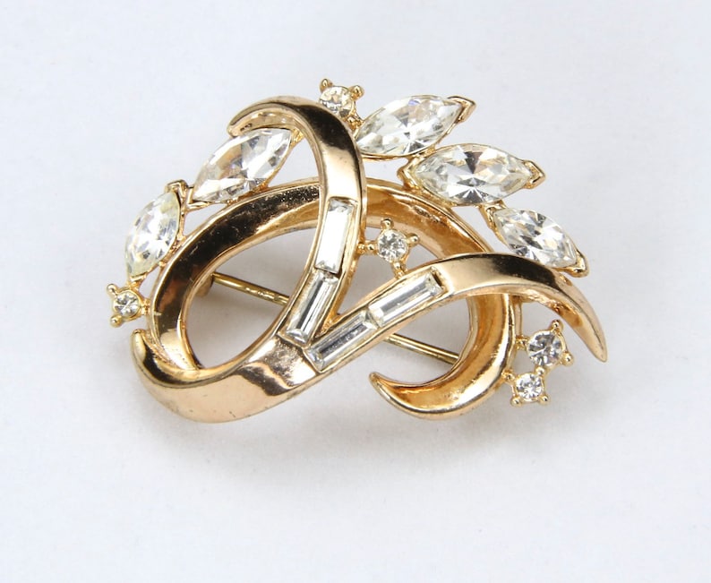May include: A gold-toned brooch with a swirling design and clear rhinestones. The brooch features a variety of rhinestone shapes, including marquise and baguette.