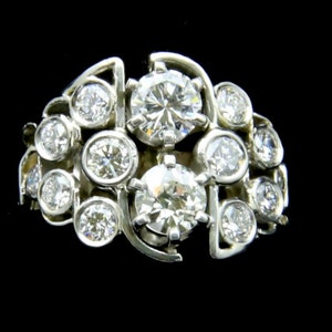 Puede incluir: Un anillo de racimo de metal plateado con m&uacute;ltiples diamantes de talla redonda. El anillo presenta un dise&ntilde;o &uacute;nico con los diamantes dispuestos en un patr&oacute;n fluido y org&aacute;nico. Los diamantes brillan sobre un fondo oscuro.