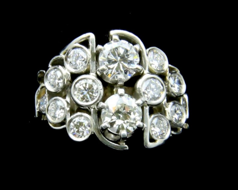 Puede incluir: Un anillo de racimo de metal plateado con m&uacute;ltiples diamantes de talla redonda. El anillo presenta un dise&ntilde;o &uacute;nico con los diamantes dispuestos en un patr&oacute;n fluido y org&aacute;nico. Los diamantes brillan sobre un fondo oscuro.