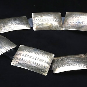 May include: A silver-toned belt with rectangular, engraved metal plates. The plates are connected by a black band, and each features a unique etched design. The belt is displayed against a dark background.