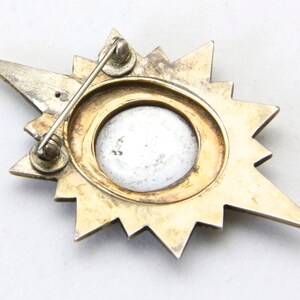 May include: Vintage starburst brooch with a silver-tone and gold-tone finish. The brooch features a central circular design with a light-colored center. The pin has a classic, elegant design.