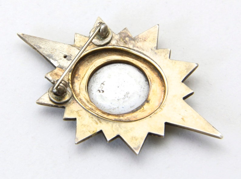 May include: Vintage starburst brooch with a silver-tone and gold-tone finish. The brooch features a central circular design with a light-colored center. The pin has a classic, elegant design.