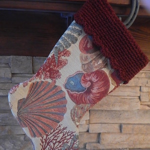 May include: A Christmas stocking with a seashell and coral pattern in red, blue, and beige. The top of the stocking is a solid burgundy knit with a scalloped edge. The stocking is hanging from a horseshoe on a stone fireplace.