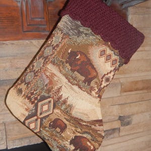 May include: A Christmas stocking featuring a woven tapestry fabric with a landscape scene of brown bison, pine trees, and geometric diamond patterns in shades of tan, brown, and cream, finished with a dark maroon crocheted cuff at the top.