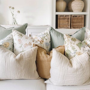 Puede incluir: Una colección de cojines decorativos en varios colores y patrones, incluyendo florales, geométricos y lisos. Los cojines están dispuestos en un sofá, creando un ambiente acogedor y agradable.