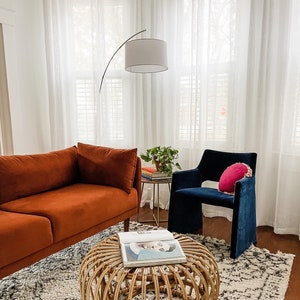 Puede incluir: Una sala de estar con un sofá naranja quemado, un sillón azul, una mesa de centro de ratán tejido y una lámpara de pie blanca con un brazo curvo. Un libro está sobre la mesa de centro y una pequeña planta está sobre una mesa auxiliar.