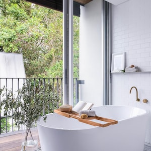 Puede incluir: Una bañera blanca independiente con una bandeja de madera y un libro encima. La bañera está en un baño con paredes de azulejos blancos y una ventana con vistas a un exuberante jardín verde.
