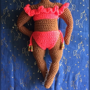 Könnte beinhalten: Häkelpuppe mit brauner Haut, die ein rosafarbenes Bikini-Oberteil und -Unterteil mit goldenen Perlen trägt. Die Puppe steht auf einem blauen Stoff mit einem goldenen Einhornmuster.