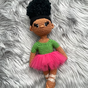 May include: A crocheted doll with brown skin, black hair, and green and pink clothing. The doll is wearing a green shirt and a pink tutu. The doll is lying on a grey fluffy surface.