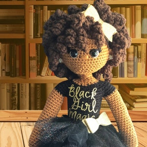 May include: Crocheted doll with brown skin, black hair, and a black dress with gold glitter. The doll is wearing a black shirt with the words "Black Girl Magic" in gold lettering. The doll is sitting on a wooden floor.