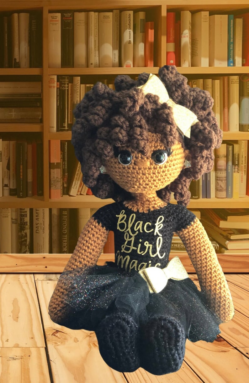 May include: Crocheted doll with brown skin, black hair, and a black dress with gold glitter. The doll is wearing a black shirt with the words "Black Girl Magic" in gold lettering. The doll is sitting on a wooden floor.