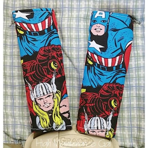 May include: Two fabric seatbelt covers featuring comic book art of Captain America, Thor, and Iron Man. The covers are primarily blue, red, and black, with white accents. The text "High Maintenance Gal" is visible at the bottom.