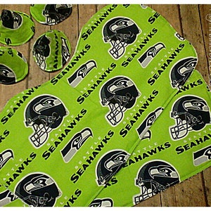 May include: Green fabric with a repeating pattern of Seattle Seahawks logos. The logos are in black, white, and gray. The fabric is folded in half and has a frayed edge.
