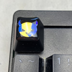 May include: A black keyboard with a single key replaced with a custom keycap. The keycap is black with a clear, glossy finish and a yellow and blue cartoon devil face graphic.