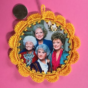 May include: A handmade, yellow crocheted ornament featuring a photo of the Golden Girls cast. The ornament has a scalloped edge and is decorated with small red beads. The background is a vibrant pink.