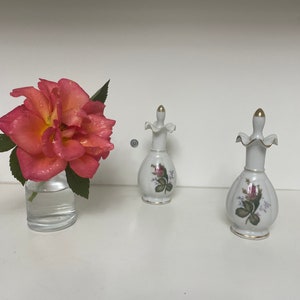 May include: Two white porcelain perfume bottles with gold accents and floral designs. The bottles have a ruffled top and a pear-shaped body. The bottles are on a white surface.