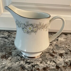 May include: White porcelain creamer with a silver trim and a floral design of white daisies with green leaves. The creamer has a rounded body and a small, round base.
