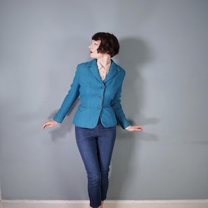 May include: A teal blue blazer with a tailored fit, featuring a three-button closure and two front pockets. The blazer is paired with dark blue denim jeans. The jacket has a textured appearance.