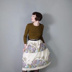 May include: A woman wearing a brown sweater and a white skirt with a floral print of fruit and baskets. She is wearing a black belt.