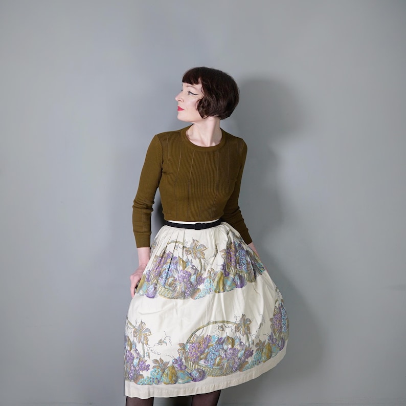 May include: A woman wearing a brown sweater and a white skirt with a floral print of fruit and baskets. She is wearing a black belt.