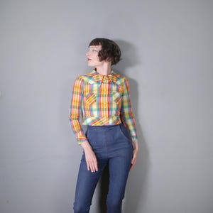 May include: A vintage, long-sleeved plaid shirt in a vibrant mix of yellow, orange, green, and blue. The shirt features a pointed collar and two chest pockets. The model is wearing the shirt with high-waisted blue jeans.